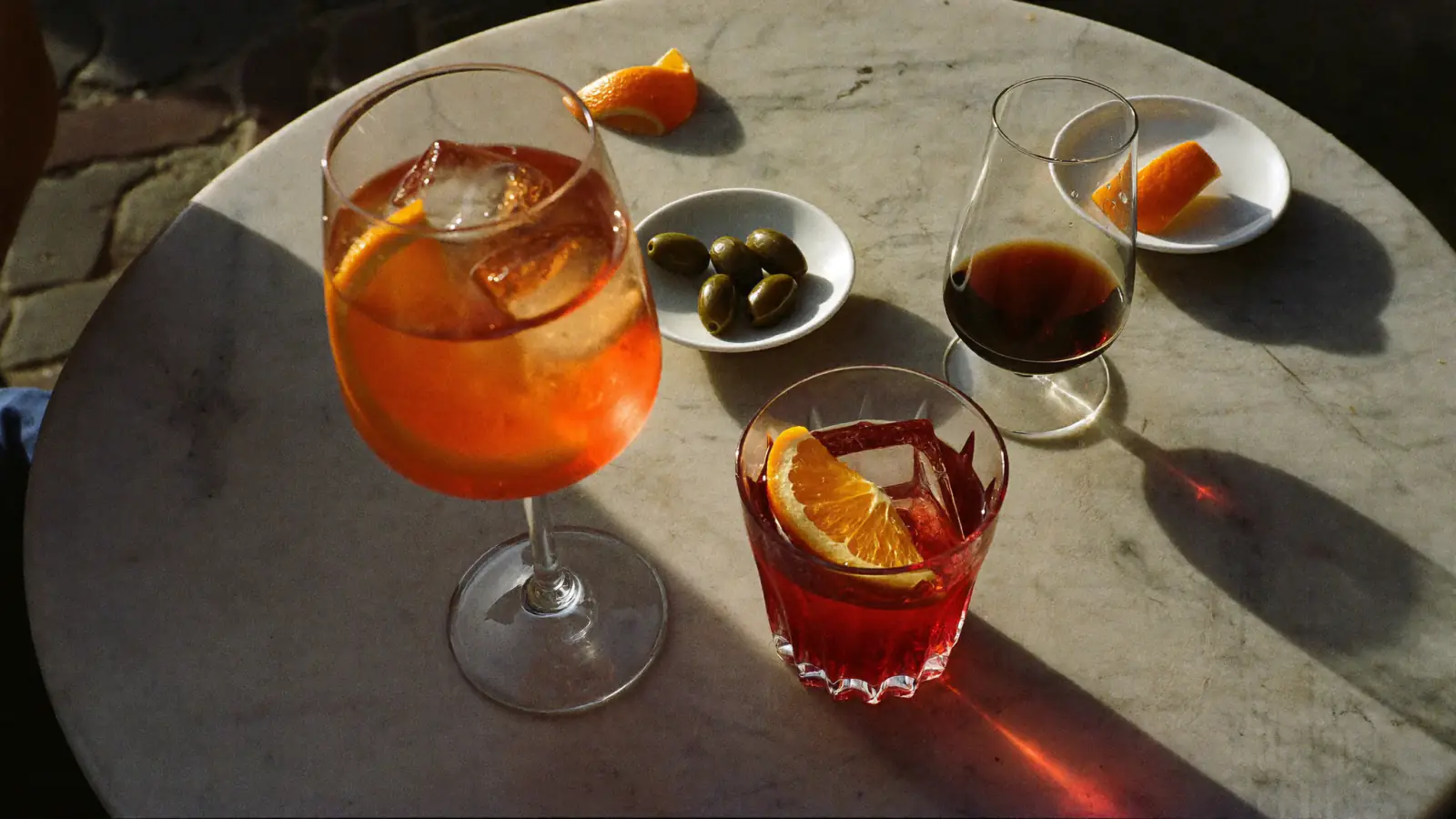 Spritz à l'Aperol, Negroni et petit verre d'amaro sur une table de café en marbre au soleil