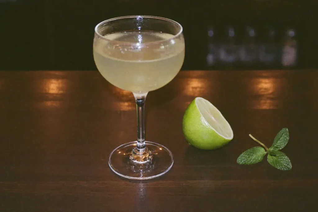 Cocktail Last Word vert pâle dans une coupe, à côté d'un demi citron vert et d'un brin de menthe