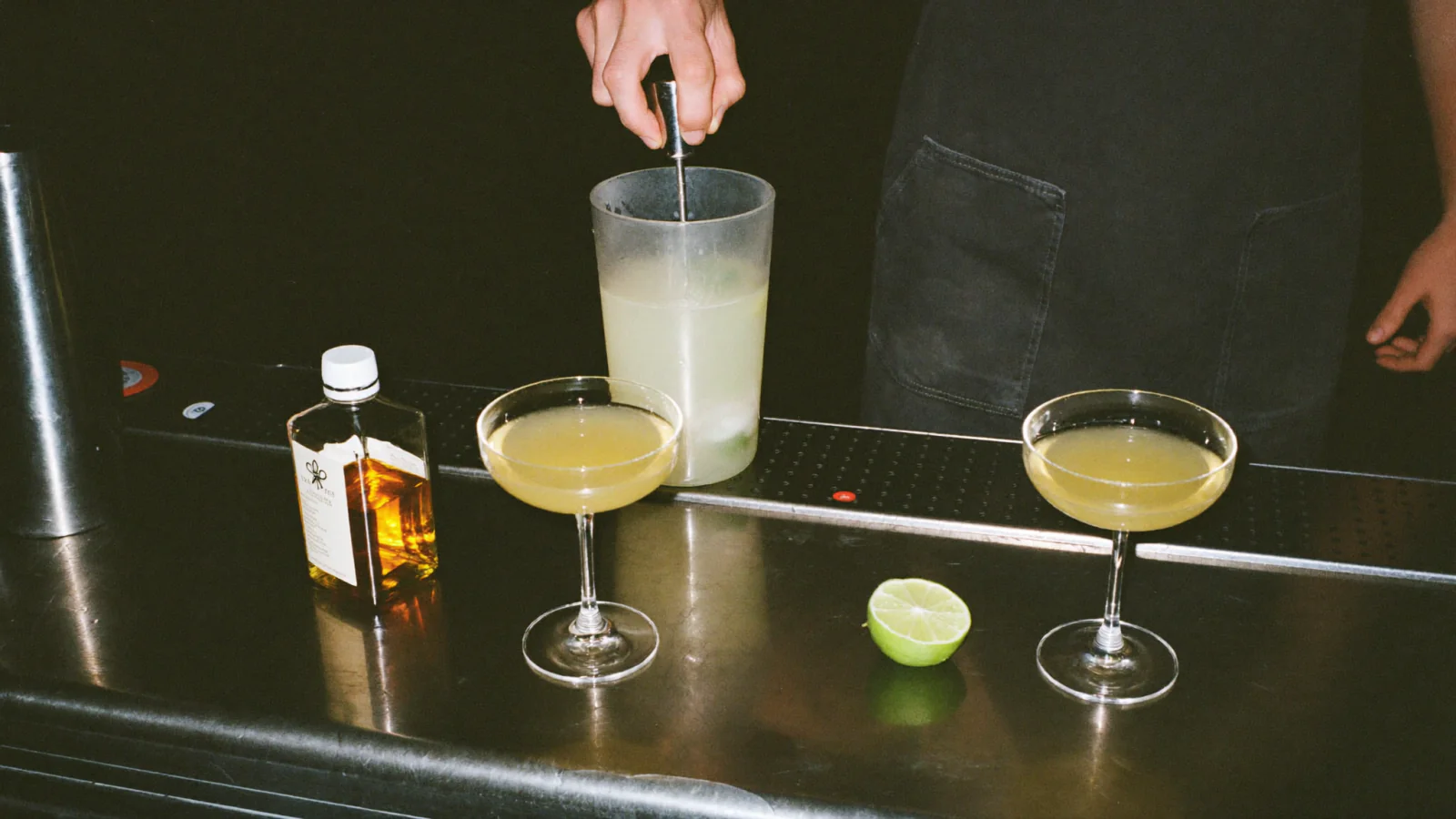 Deux cocktails servis en coupe sur un comptoir de bar, à côté d'un verre à mélange