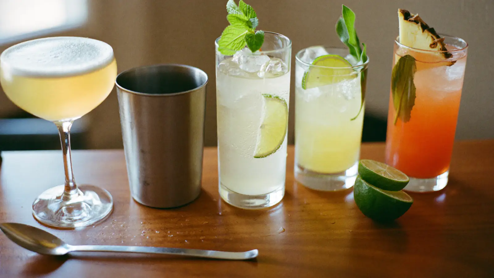 Cinq cocktails de familles différentes alignés sur un comptoir en bois : un sour en coupe, deux long drinks pétillants, un cocktail tiki et un shaker