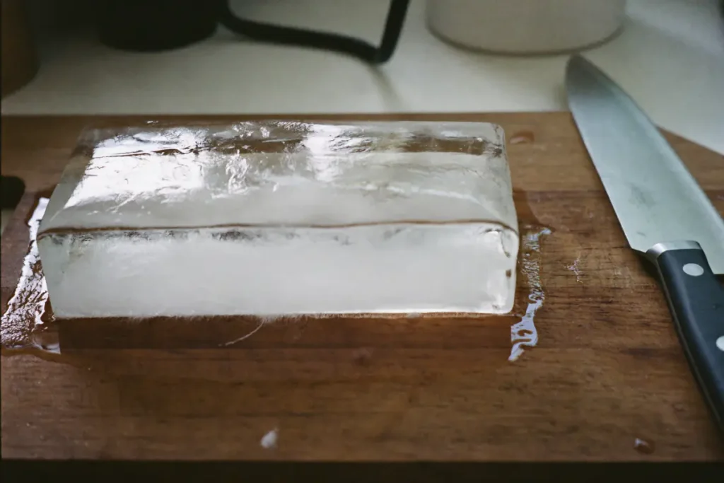 Bloc de glace maison démoulé sur une planche, transparent en haut et trouble en bas, à côté d'un couteau
