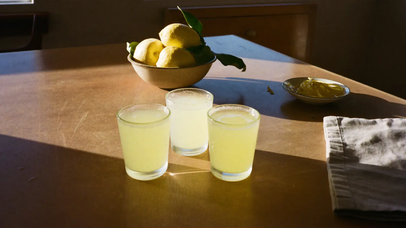 Trois petits verres de limoncello maison sur une table en bois, avec un bol de citrons et des zestes