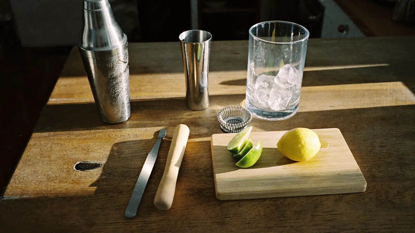 Matériel de bar à cocktail sur un plan de travail en bois : shaker, doseur, passoire, pilon, verre à mélange et agrumes
