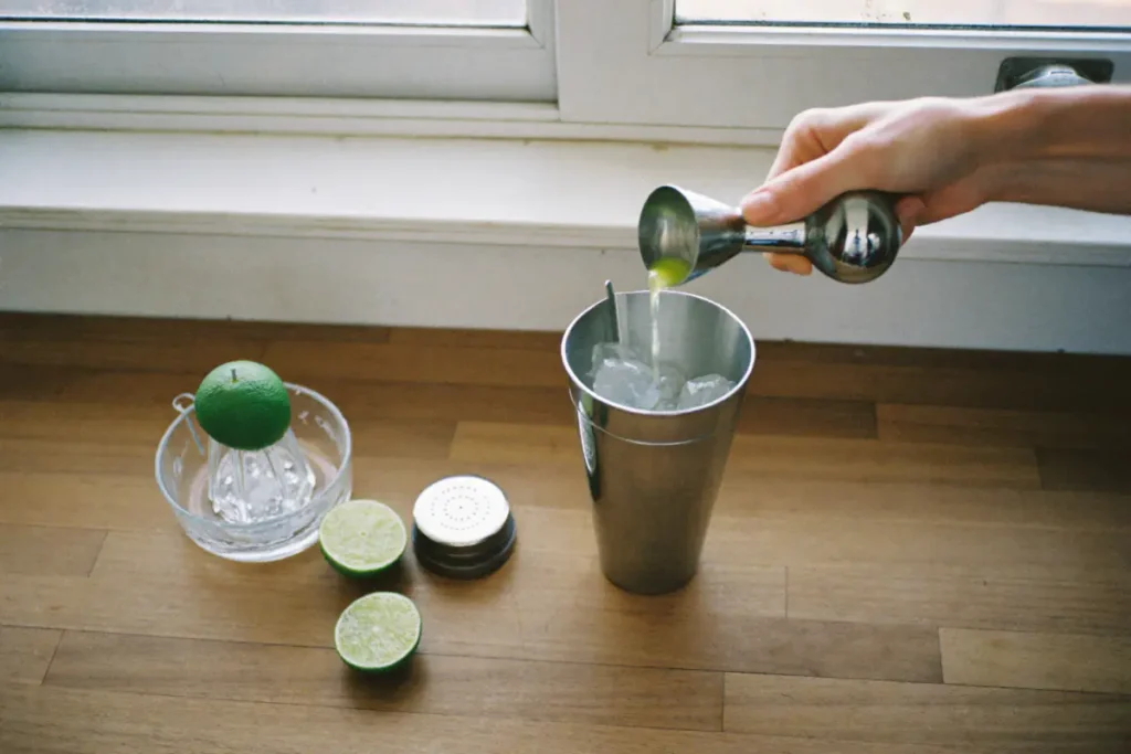 Jus de citron vert versé au doseur dans la timbale d'un shaker remplie de glace, à côté d'un presse-agrumes