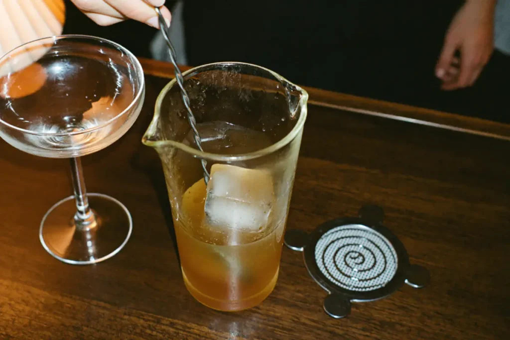 Cocktail ambré remué à la cuillère de bar dans un verre à mélange, à côté d'une coupe vide