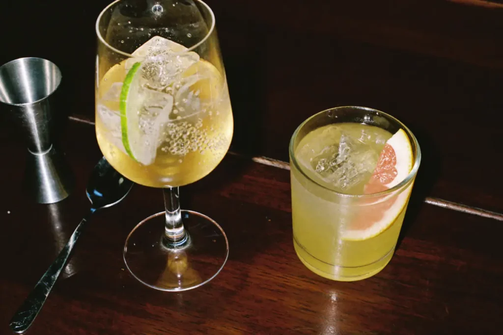 Un Lillet tonic au citron vert dans un verre à vin et un cocktail jaune pâle au pamplemousse sur un bar en bois