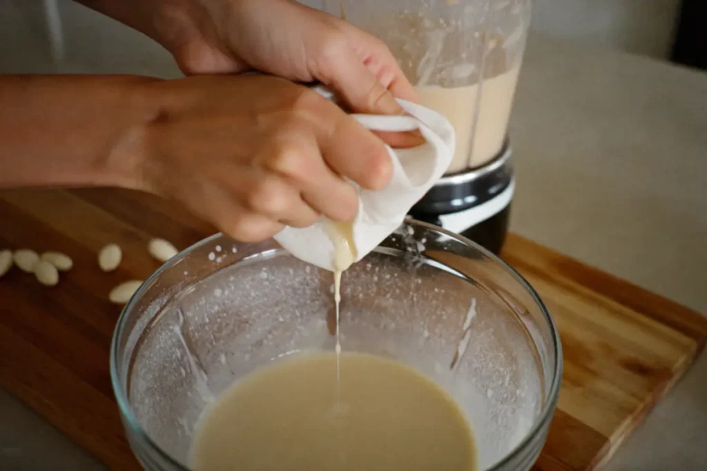 Lait d'amande pressé à travers une étamine au-dessus d'un saladier, blender en arrière-plan