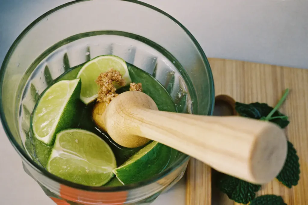 Pilon en bois qui écrase des quartiers de citron vert et du sucre de canne au fond d'un verre, feuilles de menthe intactes à côté