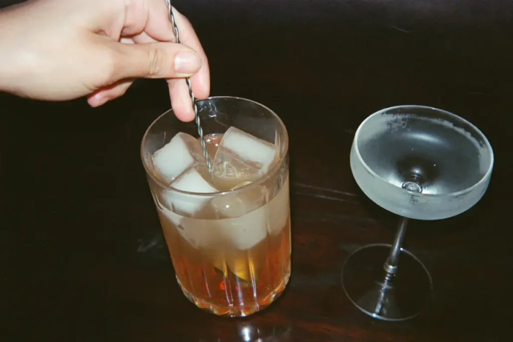 Cocktail ambré remué à la cuillère de bar sur de gros glaçons, à côté d'une coupe givrée