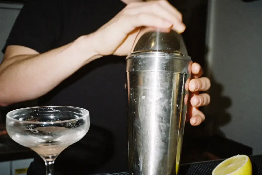 Mains qui tiennent un shaker en inox givré, à côté d'une coupe à cocktail et d'un demi-citron